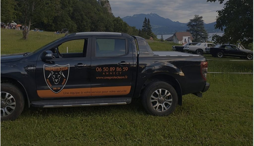 Un camion de sécurité arborant un logo et des coordonnées est stationné sur un terrain herbeux près d'un lac paisible, avec des montagnes en arrière-plan et des voitures de collection à proximité.