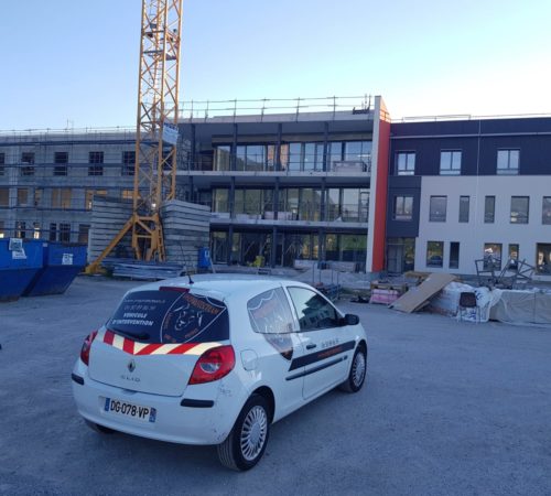 Une voiture de sécurité blanche est stationnée sur un chantier. Une grue se dresse à côté d'un bâtiment en construction, entouré d'échafaudages. Le ciel est dégagé.