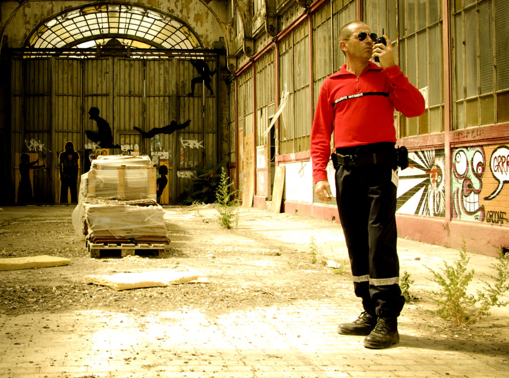 Un homme en uniforme rouge se tient dans un entrepôt abandonné, un talkie-walkie à la main. La lumière du soleil inonde la pièce, illuminant les graffitis et les œuvres d'art sur les murs.